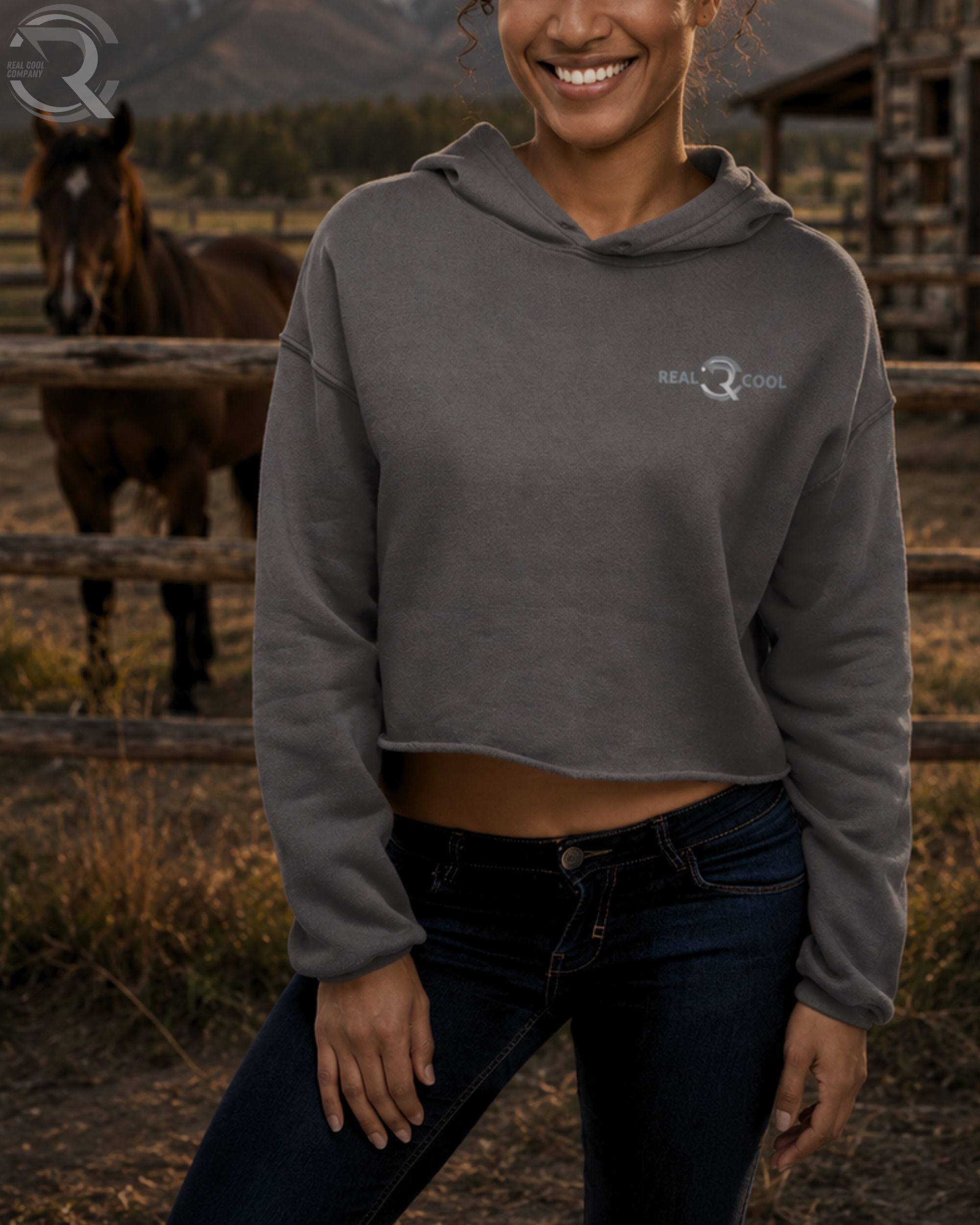 Woman wearing a gray hoodie with a logo, standing in a rustic outdoor setting with a horse in the background.